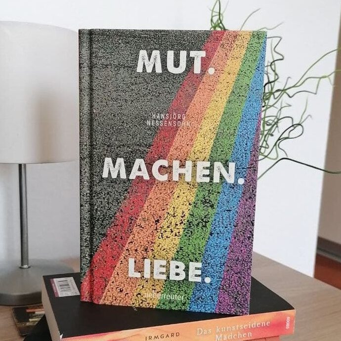 Das Buches "Mut Machen Liebe" steht auf einem Nachtschrank. Das Cover mit Namen und regenbogenfarbenen Streifen darauf ist zu erkennen.