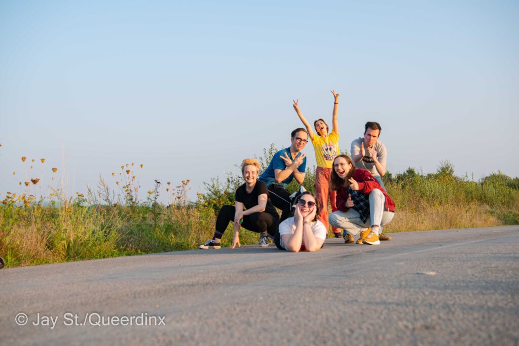 Die sechs ursprünglichen Queerdinx-Redaktionsmitglieder posen auf einem Feldweg vor blauem Abendhimmel.