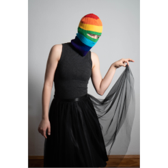 Eine Person steht vor weißem Hintergrund und hebt ihren Rüschenrock mit der rechten Hand nach oben. Die Person schaut in Richtung des angehobenen Rockes und trägt eine Regenbogen-Balaklava.
