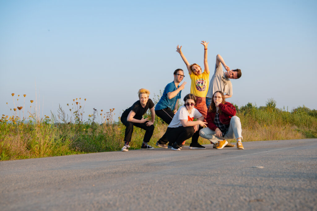 Die sechs ursprünglichen Queerdinx-Redaktionsmitglieder posen auf einem Feldweg vor blauem Abendhimmel.