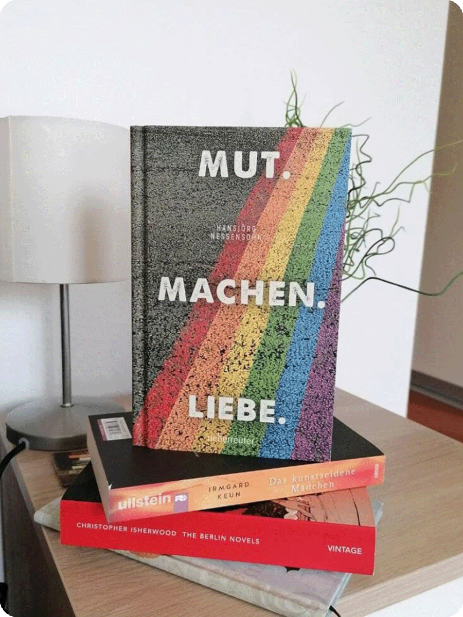 Das Buch "Mut. Machen. Liebe." steht auf einem Stapel anderer Bücher auf einem Nachttisch. Das Cover des Buches ist der Schriftzug auf einem Foto einer regenbogenfarbenen Straße.