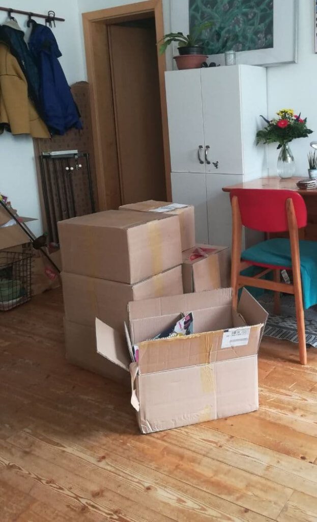 In einem Wohnzimmer stehen vor einem Tisch mit rotem Stuhl auf einem Holzboden mehrere Pappkisten aufeinandergestapelt. Die vorderste ist bereits geöffnet und man kann eine Ausgabe "Stolz" erkennen. 