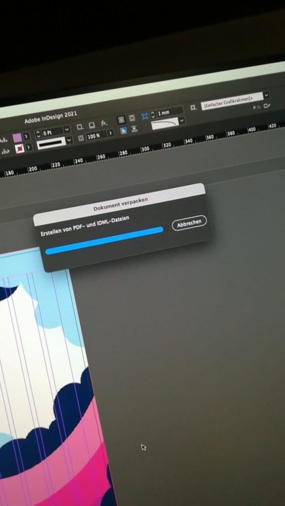 Ein abfotografierter Bildschirm ist zu sehen. Darauf erkennt man das Programm "InDesign" welches geöffnet ist. Ein Dialogfeld ist offen mit einem fast fertigen Ladebalken. Darüber steht "Dokument verpacken". 