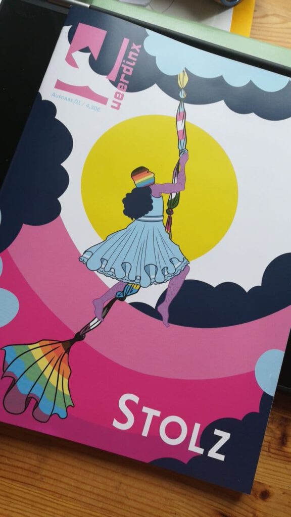 Eine Ausgabe Stolz liegt auf einem Holztisch. Auf dem Cover:  In einer blau-dunkelblauen Wolkenlandschaft mit pinken Bodenelementen schwingt sich eine Person vor einer gelben Sonne an einem aus Pride-Flaggen zusammengeknüpften Seil in die Höhe. Die Person hat helllila Haut und trägt ein blaues Rüschenkleid sowie eine Regenbogen-Balaklava. Sie klettert an dem Seil in die Höhe. Links oben ist das Logo von Queerdinx eingebettet und rechts steht “STOLZ” in Großbuchstaben.