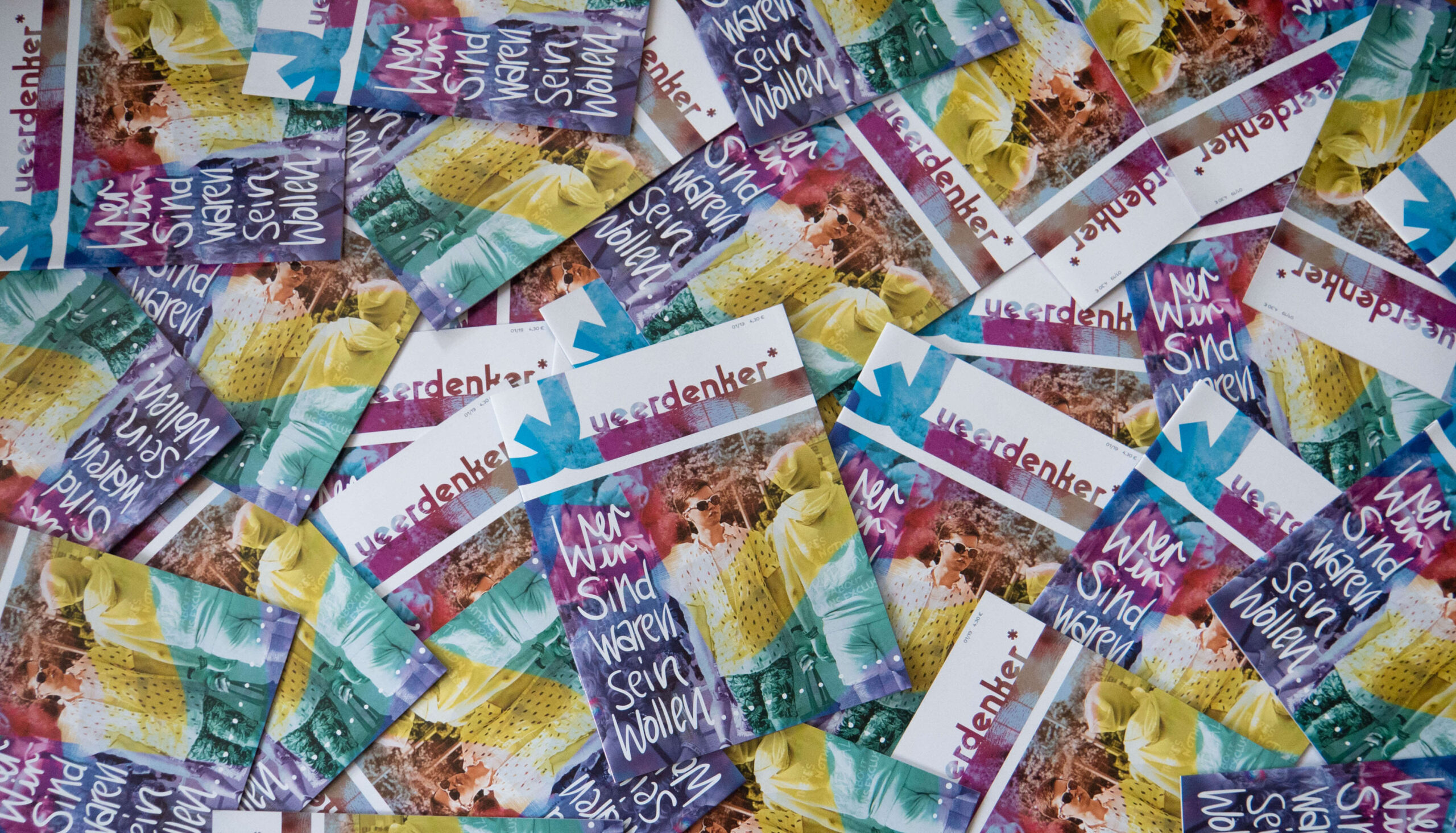 Viele Magazine liegen übereinander wild verteilt. Auf dem Cover des Magazins ist der Schriftzug "Queerdenker*" zu lesen sowie in Schreibschrift "Wer wir sind waren sein wollen .....". In  Regenbogenfarben bearbeitet ist eine Person im Vordergrund einer Fotografie zu sehen. Die Person trägt ein Hemd und eine Sonnenbrille und ist umgeben von anderen Menschen. 